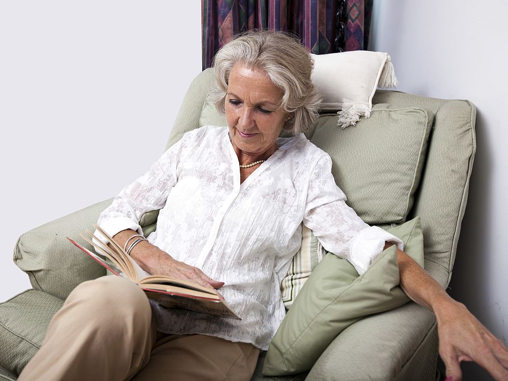 A grey-haired white woman sits in a comfy-looking armchair reading a book. She wears a plain white blouse and beige trousers and has a string of pearls around her neck. 