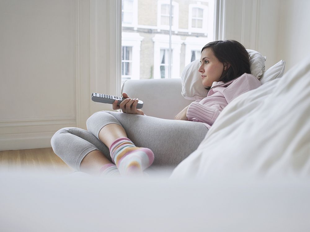 A young white woman lies cozily on a white sofa. She faces to the left of the photo and has a remote in her hand, also pointing to the left. She wears grey leggings and a pink shirt.