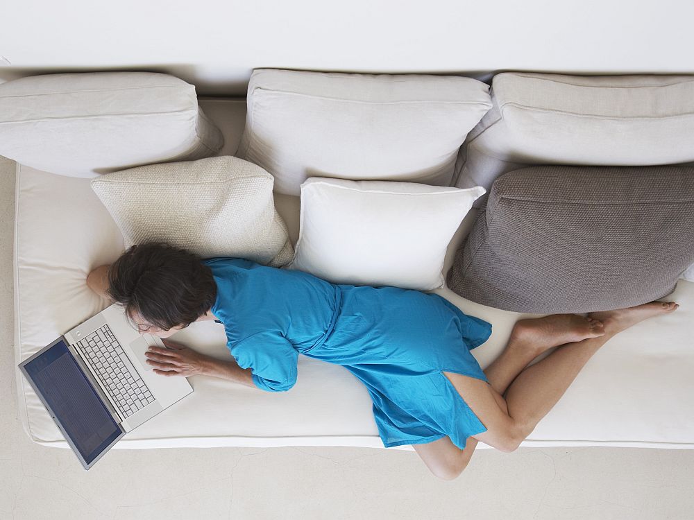 Seen from above, a white or Asian woman sprawls on a white sofa, looking at a laptop screen. She wears a simple blue dress and has bare feet.