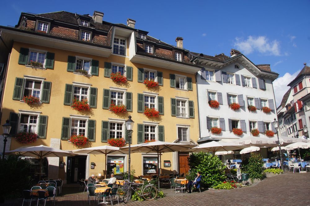 Left, a large building with about 6 stories, painted yellow with green shutters on the windows, 6 windows across each story. Red flowers in flower boxes under most of the windows. Next to it, right, another building, same height, but only four windows wide. This one is painted white with grey shutters and also has red flowers in window boxes. In front of them: tables outside with umbrellas above them.