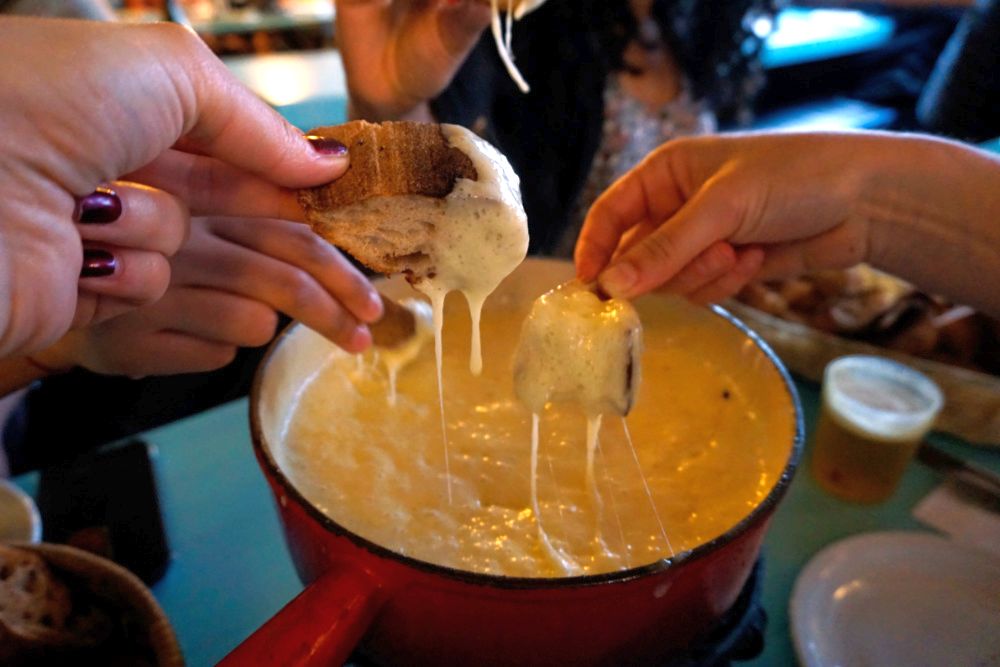 A pot of fondue, and three hands dipping chunks of break into it.