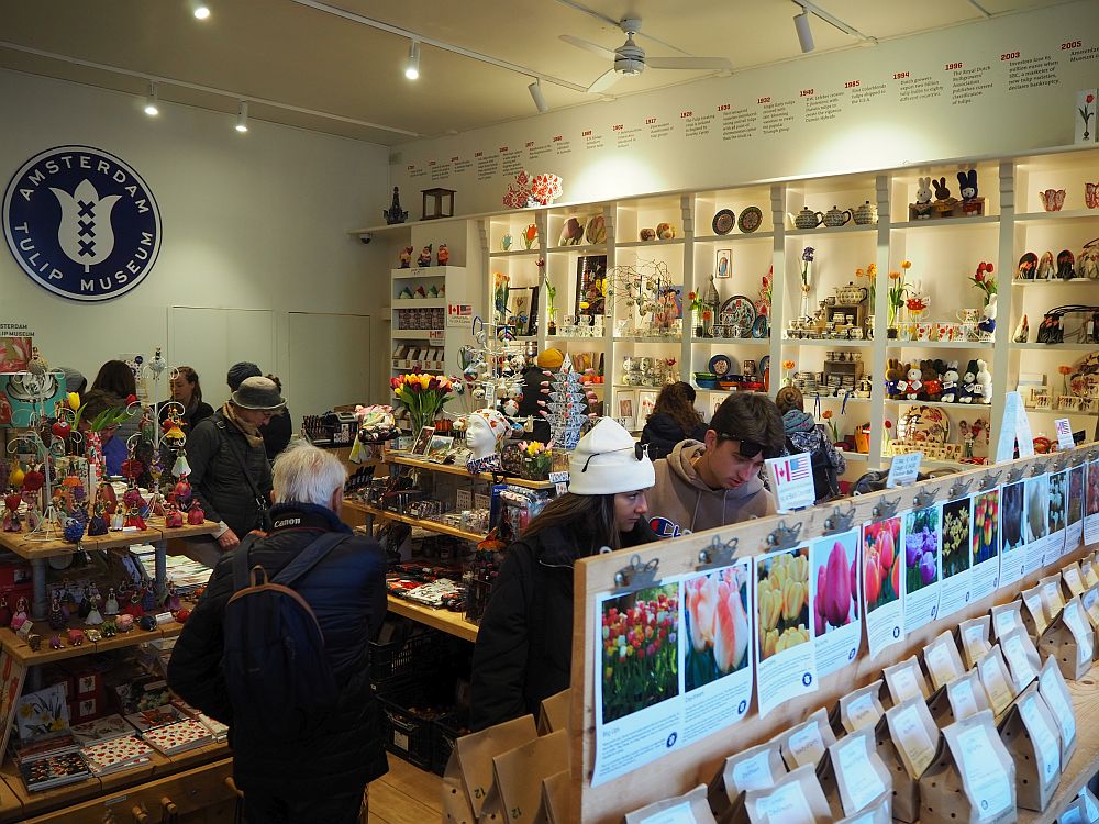 The shop in the Amsterdam Tulip Museum, seen from slightly above, filled with shelves and tables and lots of people among them. Bags of tulips are visible in the foreground and lots of brightly-colored objects cover the shelves and tables. 