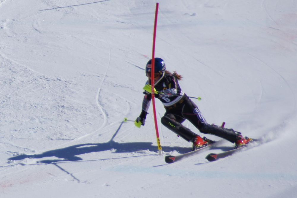 Shiffrin, in a black ski suit, a black helmet and red ski boots, swooshes around a red pole. She is leaning and clearly moving very fast.