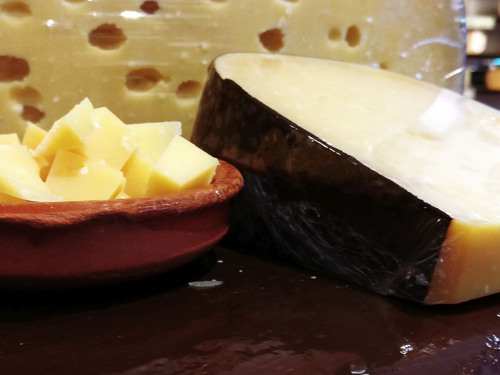 A close-up of, on the right, a wedge of cheese with a black wax out shell and, on the left, small cubes of cheese in a shallow bowl. Behind is a larger cheese with a lot of holes.