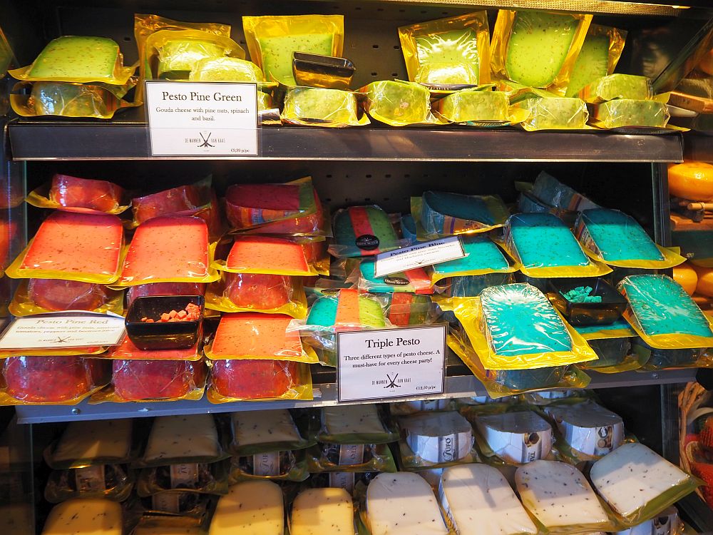 piles of shrink-wrapped cheese wedges: yellow on the tops shelf with a sign reading Pesto Pine Green. Orange and bluish green on the middle shelf, with only one sign visible that says Triple Pesto. The cheeses on the bottom shelf are a more normal color.