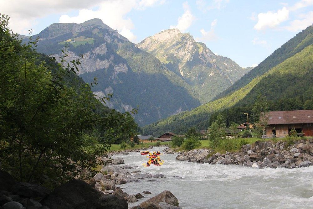 A river in the center of the photo cuts through between two mountains, with more mountains behind. In the foreground, rubber rafts on the river. A small house is partially visible on the riverbank on the right.
