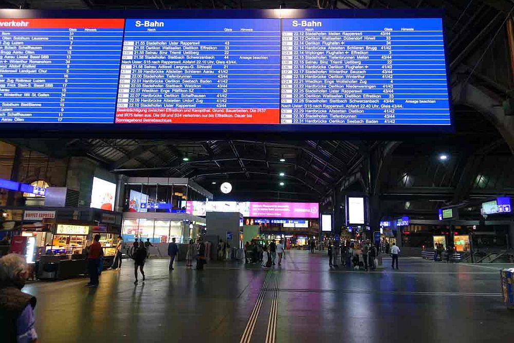 A view across a large train station: dark ceiling and floor, with lighted shops along the sides, as well as signs. In the foreground, a large, blue electronic sign showing a list of trains, times and tracks.