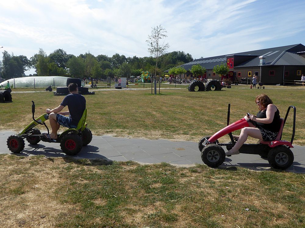 In the foreground, two people (adults) ride some sort of four-wheeled pedaled vehicles. In the background a bouncy structure and various other play structures can be seen, as well as a very large barn.