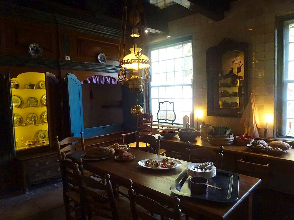 This interior is indistinguishable from a real, full-sized kitchen. Wood table with chairs around it and dishes on it. A counter behind it, under two windows, with various items for food preparation. A lamp lights it all from above in a yellowish color. In the back wall a cabinet is open, showing decorative china, also lit in yellow. Next to that, an open bedstee (a cabinet bed).