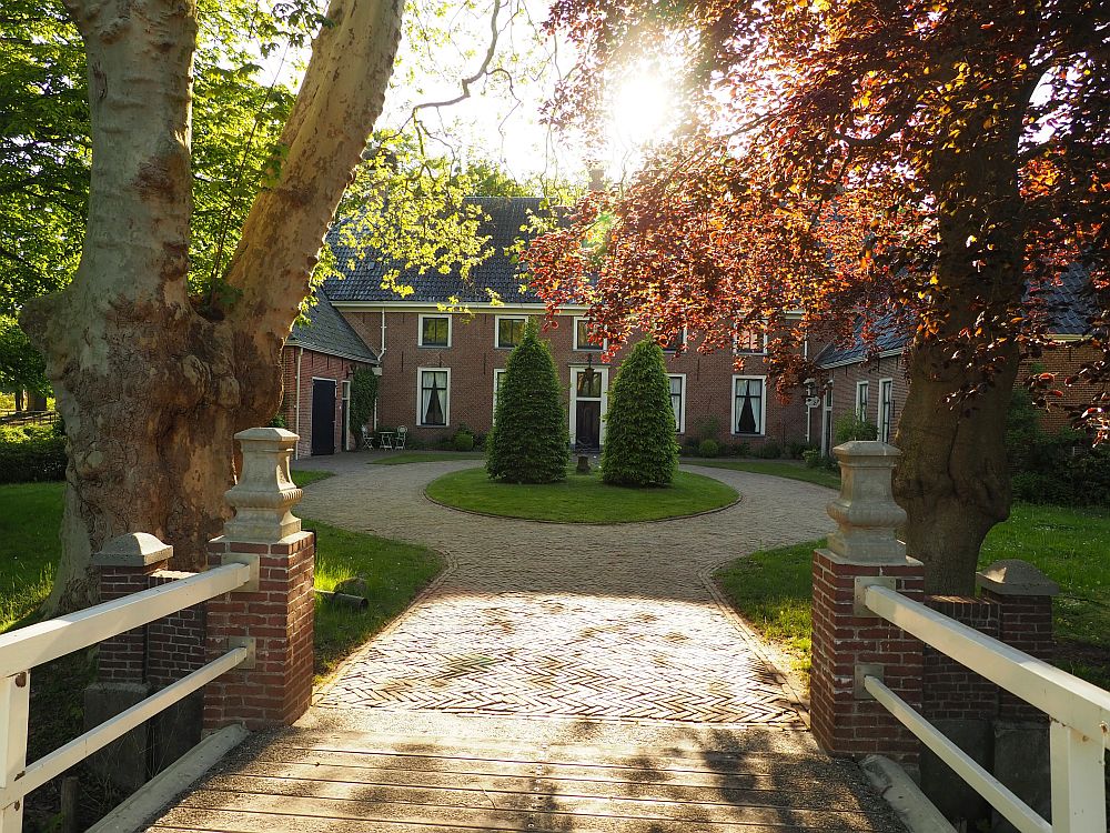 Looking straight down the entrance to the estate: the path straight ahead, then splitting into a ring. In the green island it forms: 2 neatly trimmed pine trees. The house is behind that: red-brick, with two rows of windows. Tall windows on the ground floor, square ones above each. Door in the center. The wings on each side of the entry are not quite symmetrical like the center house is.