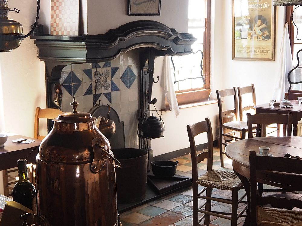 A chimney with blue and white tiles at its back wall (presumably Delft), a large kettle hangs in its center and another to the side. The mantel is dark would and shaped in a curve at the center, but otherwise relatively plain. In front of the fireplace is a round table, partially visible, with simple chairs around it and a teacup and saucer in front of one of the places. Beyond that, a window and a square table with chairs. IN the foreground, a big copper container of some sort: for coffee, perhaps.