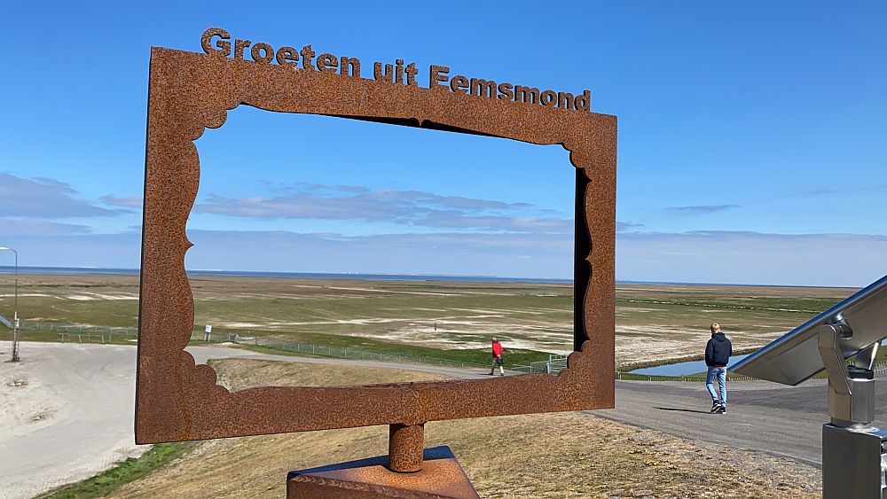 A large rectangular rusty steel frame with the words "Groeten uit Eemsmond" frames a very flat scene of grass and sand into the distance, where a line of blue sea is visible, with a barely perceptible rise of land beyond it. A few people walk toward the sea along a dirt path next to the frame.