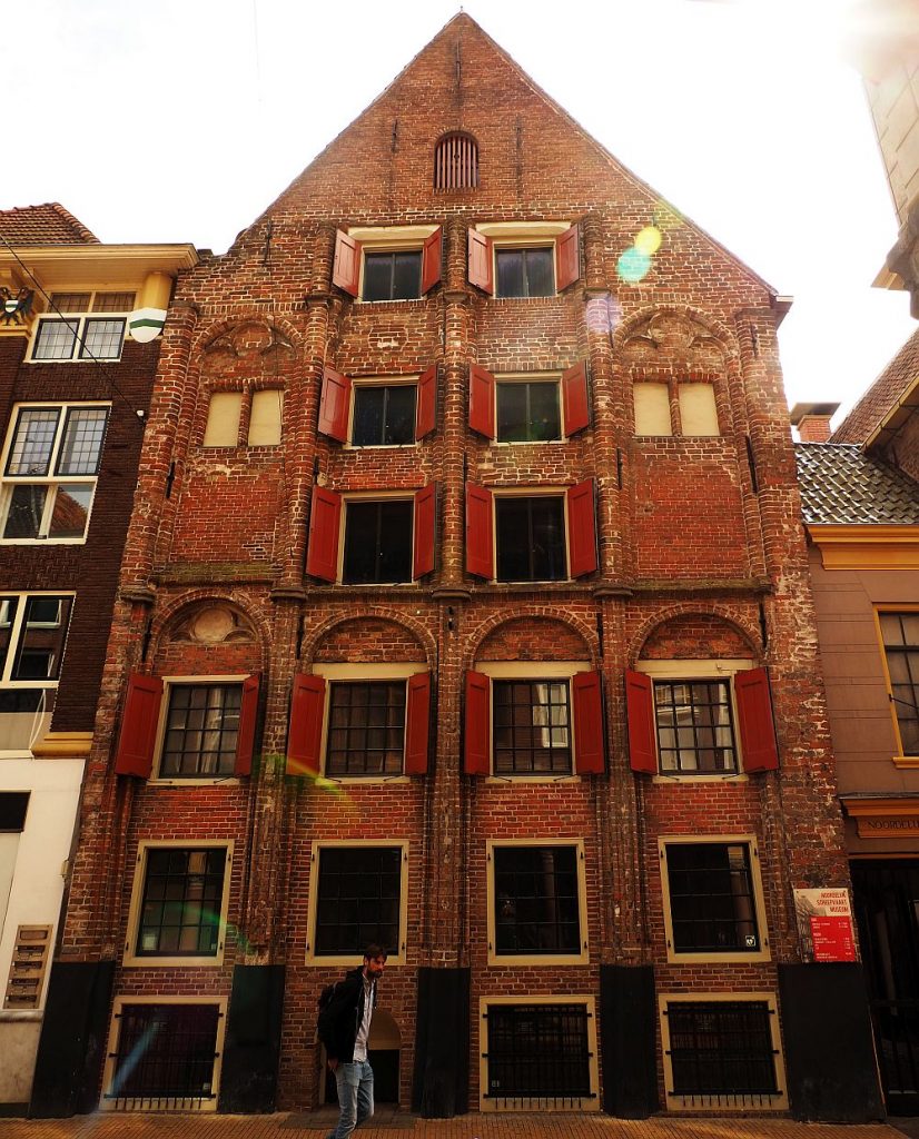 A very old brick building in a row. It appears to be six or seven stories high. Some of the upper windows have red shutters. Most of the floors have 4 windows across.