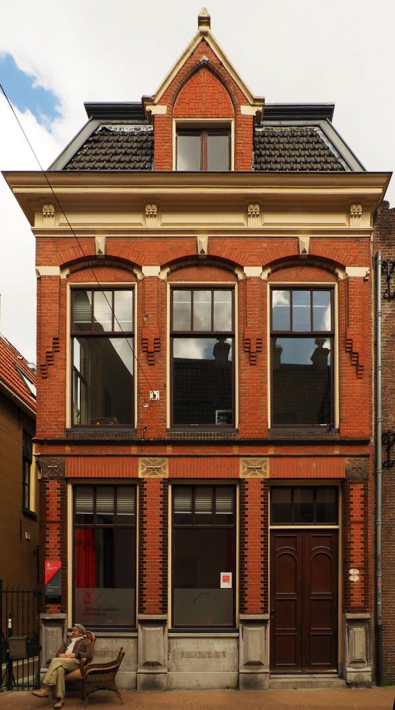 A two story row house. The grown floor has two tall windows and a door. The upper floor has three tall windows across. Above that, at the attic level, is one single dormer window. The building is red brick, with a wooden door. There are brick decorations between the upper windows and dark bricks along the edges of the lower windows. 