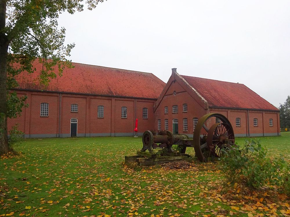 Red-brick industrial buildins in the background, about two stories tall, with red roofs. In the foreground, some sort of machinery, with a big wheel or gear. 