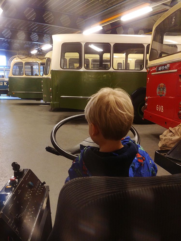 In the background, a row of buses, only the back ends visible in this photo. In the foreground, a blond child with his back to the camera. He is sitting in a driver's chair and pretending to steer a very large steering wheel. 