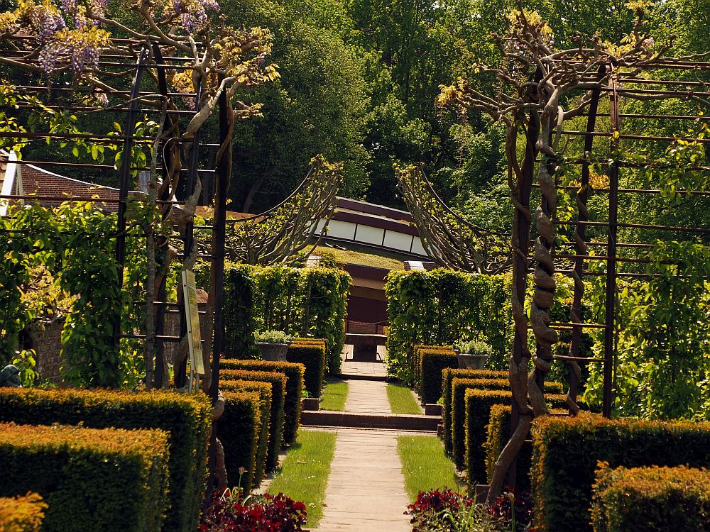 The path goes straight ahead. On either side are very neatly trimmed hedges ending at the path. An arbor on each side ends at the path as well: each has some sort of vine or tree trained around it. Behind are more, but taller hedges, and beyond them is a bit of grass rising up to the peak of the roof, which does not appear to be very high.