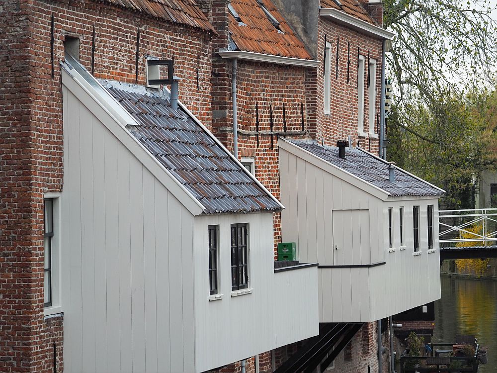 Three old buildings on the edge of a canal: all are red brick, old and uneven. Two of them have white-painted wood extensions, each with a slanted roof, hanging over the water. Each is the size of a rather small room and has several windows. 