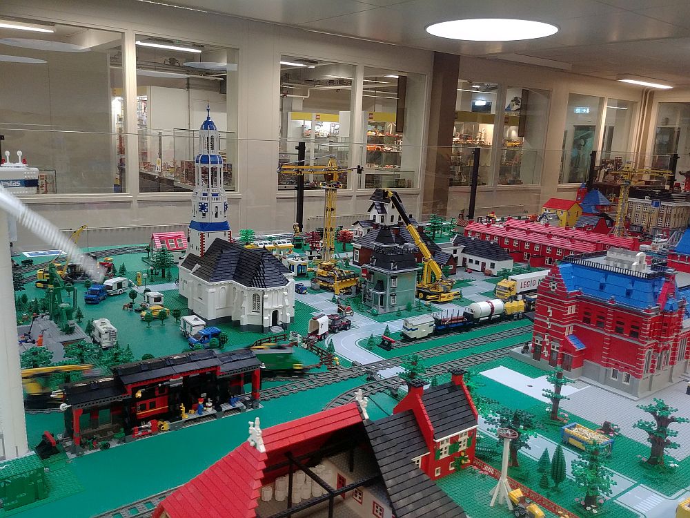 A large table covered with Lego constructions: a church, various other buildings, cars, trucks, trains, etc. Through windows in the back, more Lego can be glimpsed. 