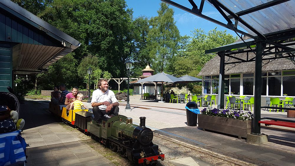 A very small train: a man sits on top of the engine. A small boy is able to sit in one of the roofless train cars, but an older child sits opposite him on top of the train car. 