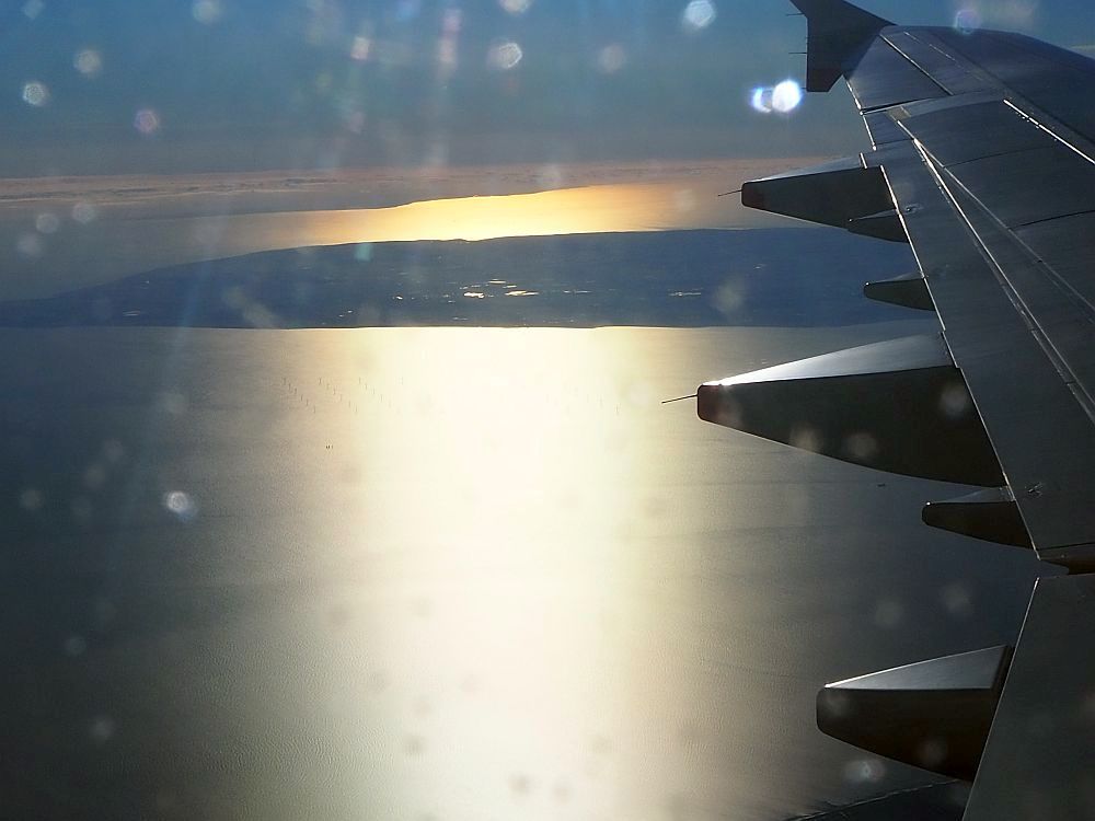 Looking out at the wing from a seat just behind the wind. Along the right side of the photo, only the back edge of the wing is visible. Below is the sea and a flat bit of land in the background. The sun makes the water shine yellow down the center. The sight is scary for a fearful flyer.