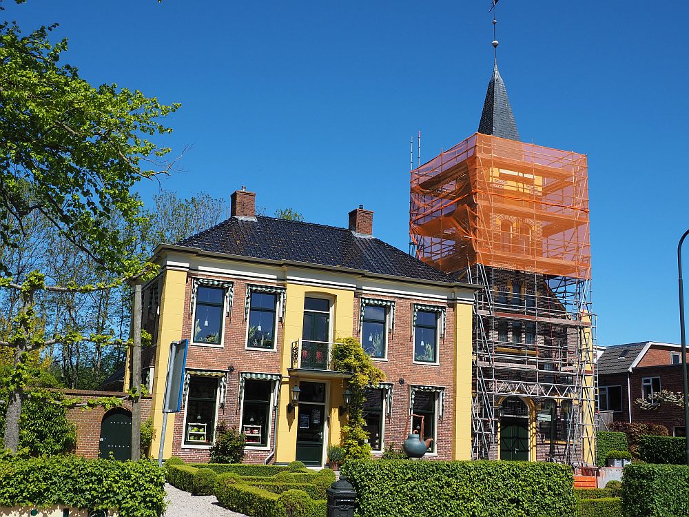 A stately-looking house, red brick and yellow plaster: symmetrical, with two stories, two doors in the center, a small balcony between them, and two windows on either side on both stories. Next to  the house is a church with a steeple, but it isn't very visible because it is covered in scaffolding.