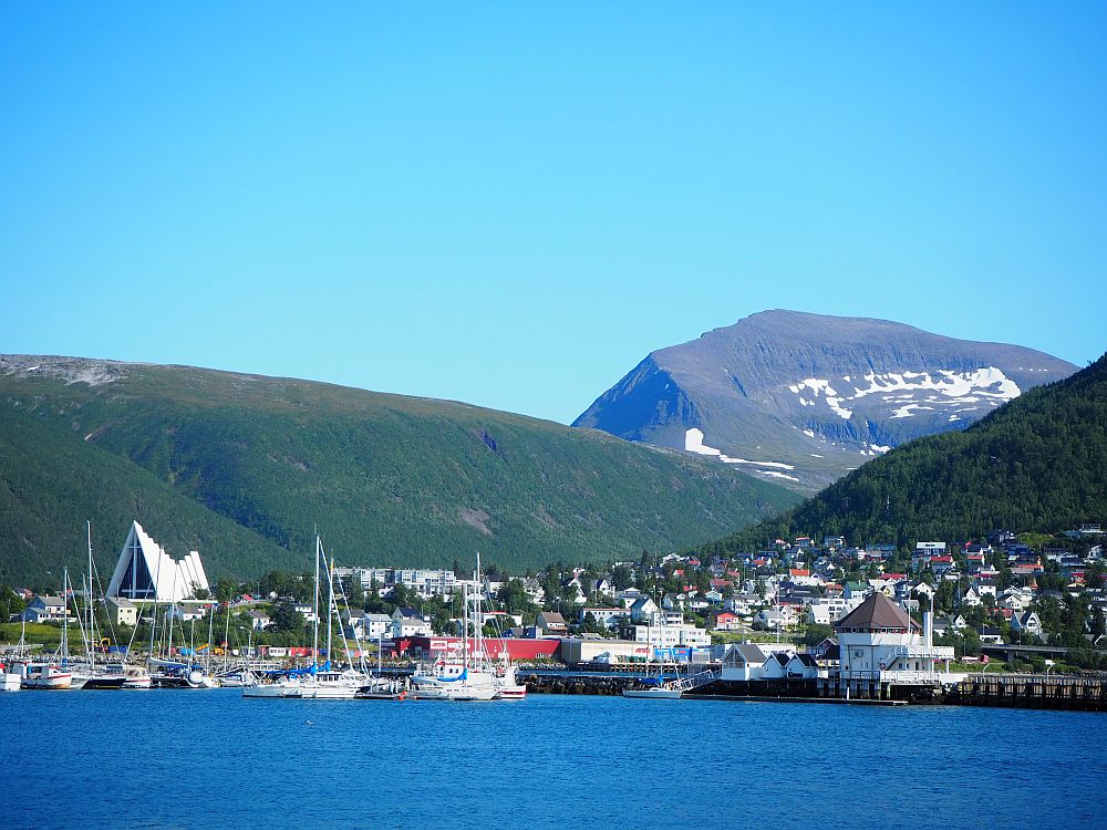 In the foreground, calm blue water. The buildings in the middle ground are mostly quite low-rise, clustered between two green-covered mountains left and right. In the background, another higher mountain, this one partly green but also with remnants of snow visible. Blue sky above.