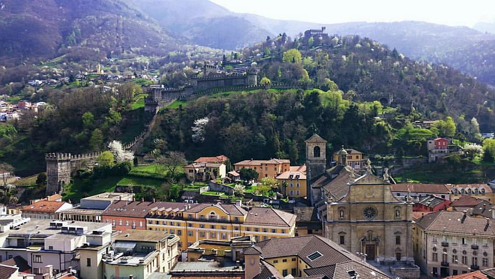 In the foreground, a cluster of buildings. Behind are two hills, one in front of other. Some castle walls are visible on both hills. 