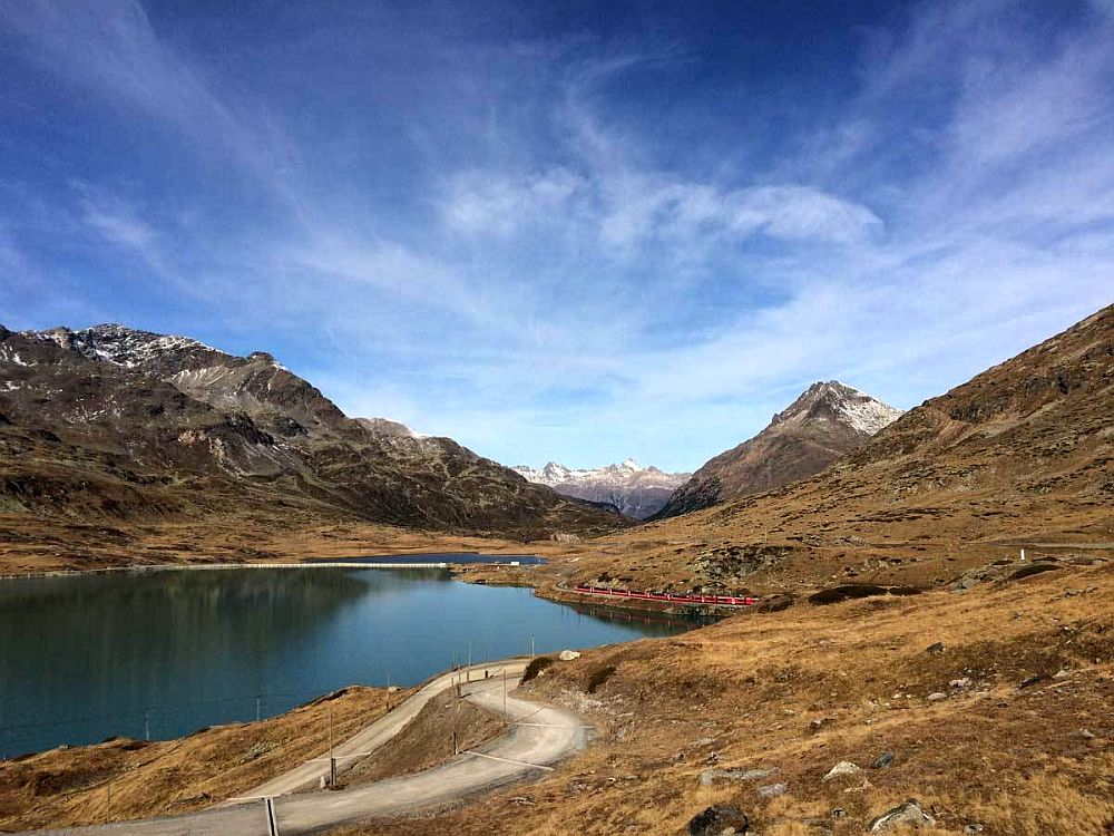 A lake on the left, with mountains behind it. Along the righthand lakeshore, a red train is visible on a track that follows the shore. More mountains in the distance.