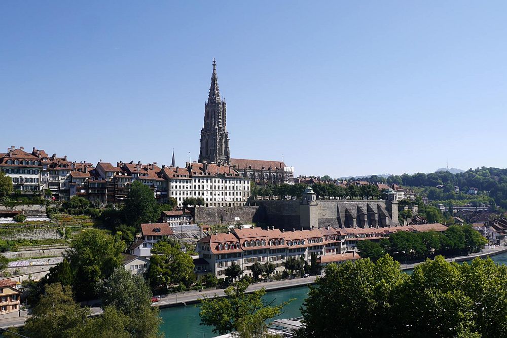 In the foreground, the bend of a river. On the other riverbank, a clustered town on the side of a hill. At the top, a church with a very tall tower. Berne is one of the UNESCO sites in Switzerland.