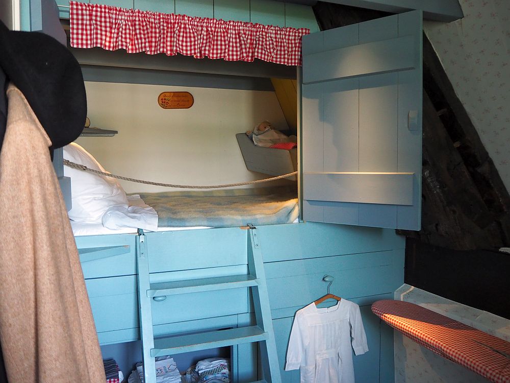 The bed is built into a wall, with a blue painted door standing open. A small ladder of just a few steps, also blue-painted, leads to the bed, which appears quite small. Under the bed are some shelves. Inside, the mattress, blanket and pillows are visible. At the foot of the bed, inside the bedstee as well, a small cradle seems to be mounted on the wall. 