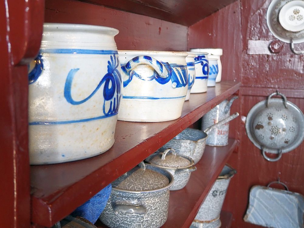 On a red-painted shelving unit, a row of simple pots: white ceramic with simple blue decorative paint. Below them, a row of enamelled metal pots, all in a gray pattern.