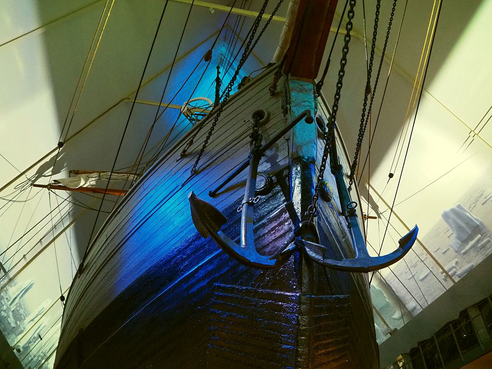 Looking up at a large pointed hull. It looks like it's made of wood planks, though that may not be so. Two large iron anchors hang from chains near the point of the bow. Beyond the boat, images are projected on the two walls of the building: a sea with floating icebergs.