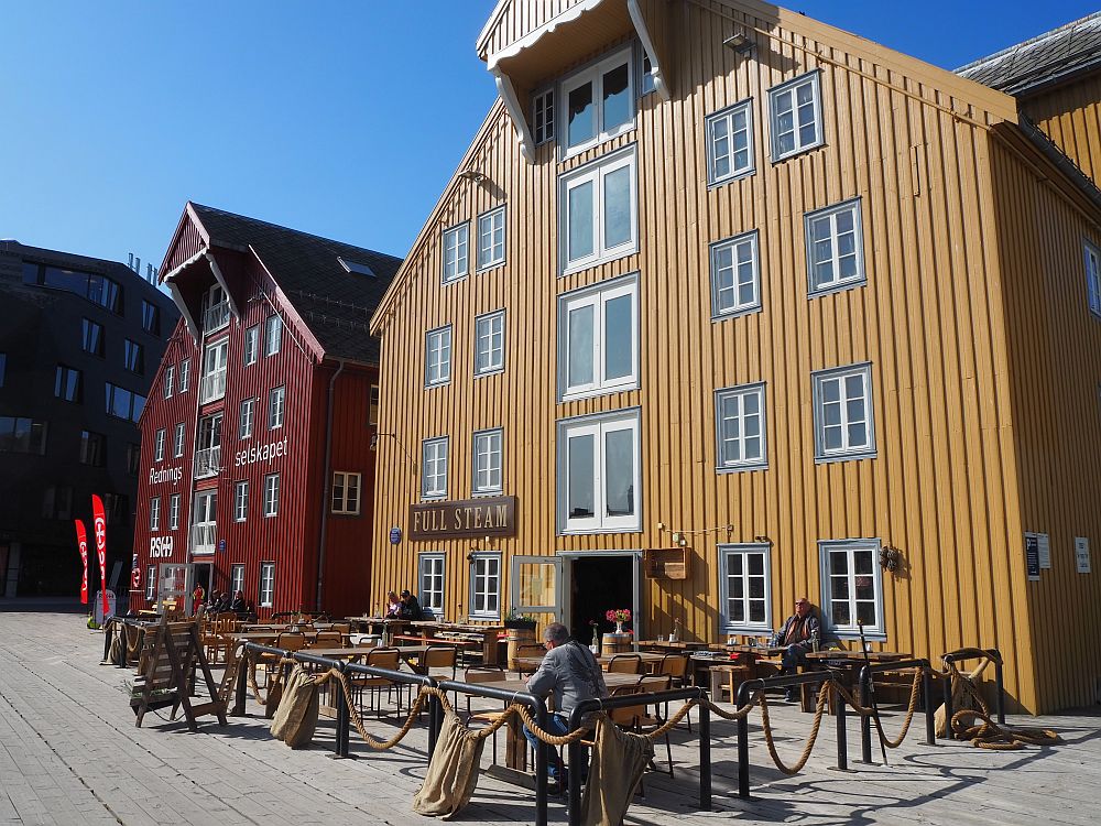 Two converted warehouses: red on the left, yellow on the right. Both are wood with vertical panels. Both have a peaked roof with a gable extending over the street (to hold a pulley, I assume) over a vertical line of big windows that were presumably originally warehouse doors. On either side of the column of bigger windows are smaller paned windows, two on each side on each of 4 stories. In front, a sidewalk cafe with a scattering of people at the tables.