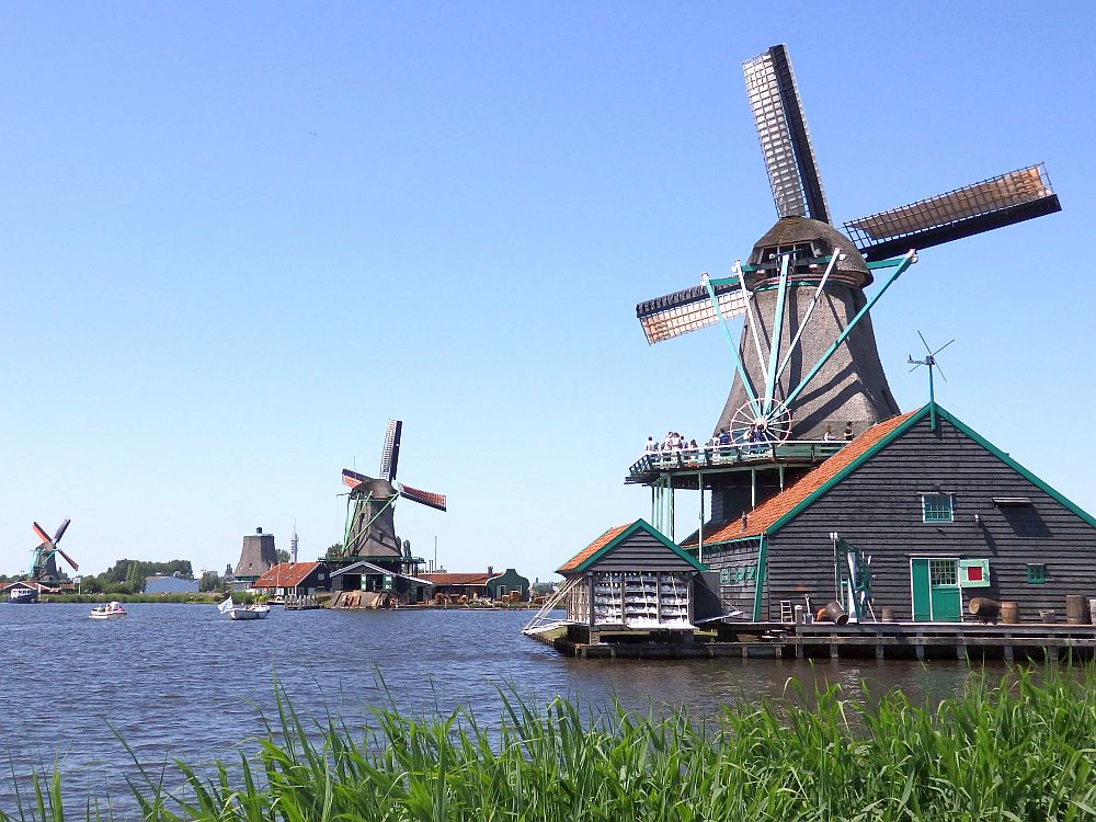 KInderdijk or Zaanse Schans? Three windmills are visible in this picture along a body of water. The nearest one seems to be six-sided and is perched on top of the roof of a house (but I think it's a workshop/factory). All are made of wood, and the house is on stilts above the water.