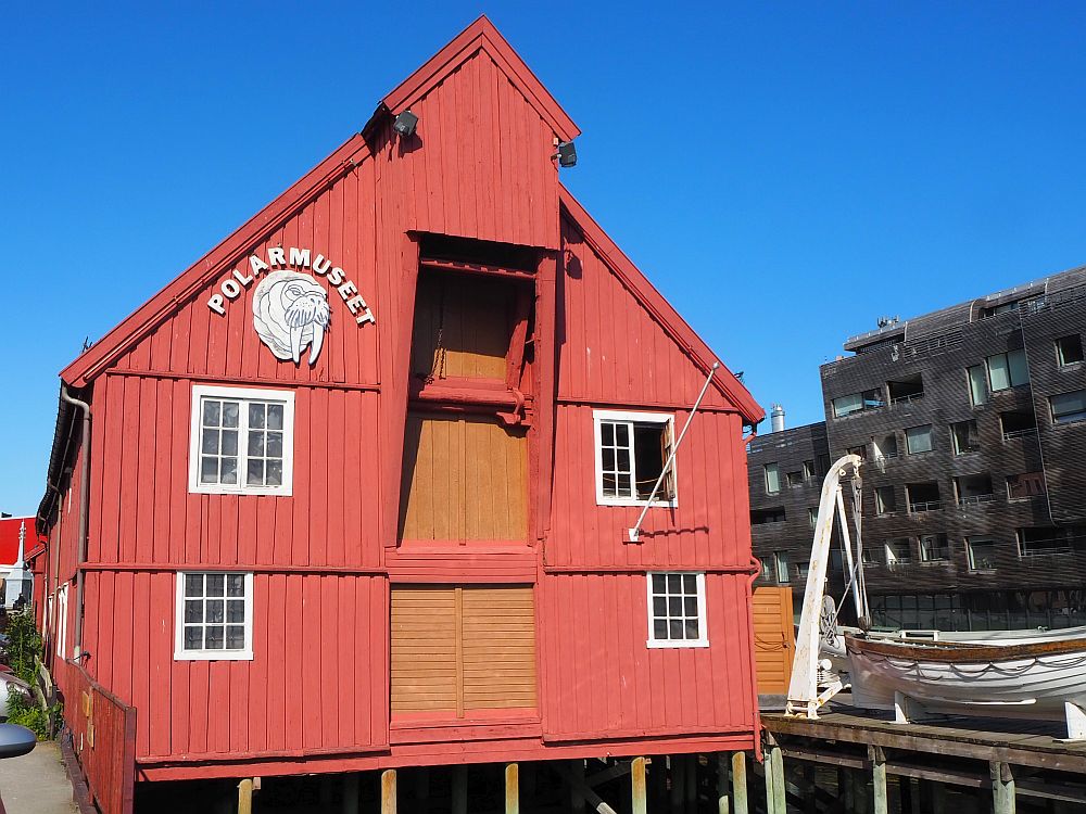 The Polar Museum in Tromso is in a former warehouse, painted red. The roof is peaked and has a gable that extends out from the flat front, presumably to hold a pulley to lift things up. There are three stories. Up the middle are big openings on each floor, covered in board now. On the ground floor and one floor up: one window on each side of the bigger wood-covered openings. The building, or at least this side of it, stands on stilts over the water. Above the two left-hand windows is a white sign: Polarmuseet, with a face of a walrus below it. 