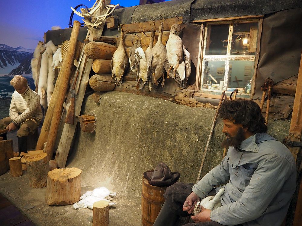 A log cabin with one small window. Hanging next to the window are a number of dead birds: geese or ducks. On the right side of the picture a man sits, and it looks like he's doing something to a dead animal, maybe a fox: perhaps skinning it. There's another lying on the ground. On the left side of the picture another man sits in front of a bunch of arctic fox furs hanging on the wall of the cabin. 