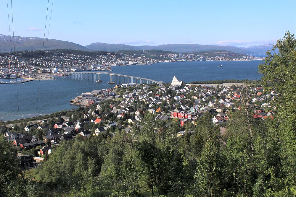 The nearer part of the city is a cluster of low buildings, with the Tromso Cathedral standing out as a bigger white building. The body of water is crossed by a long bridge, and beyond the rest of the city sprawls along the water, also low-rise. Beyond that: mountains.