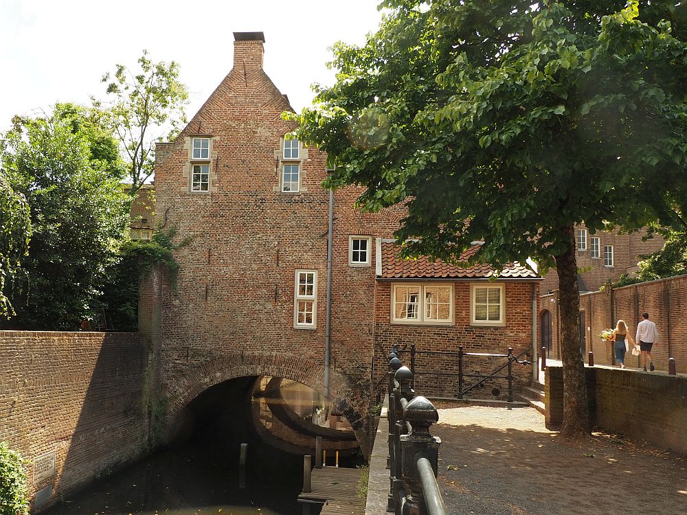 Another canal-like stream flowing under an archway with a brick bulding on top of the arch. This building is two stories tall. On the left is a brick wall with treetops visible beyond it. On the right is a walkway with a railing along the stream. Further to the right at the edge of the picture is a road, and two people walk down the road.