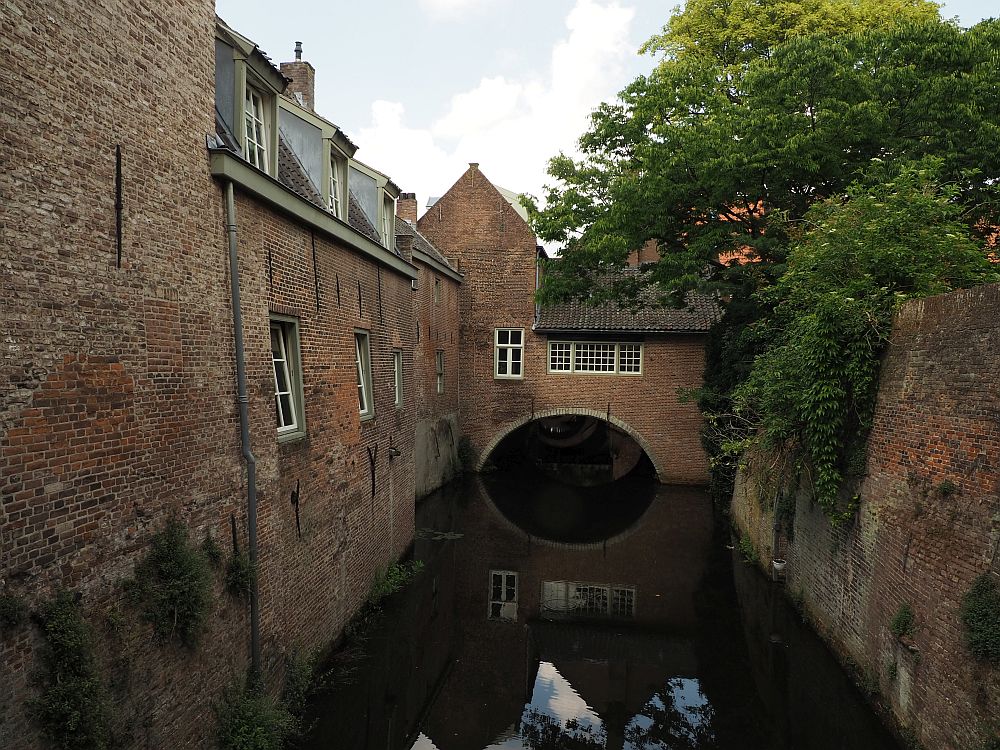 A small, straight canal down the middle of the photo disappears under and archway with a small brick building spanning the archway. A plain brick wall on the right, a tree top showing over it and dangling down its side. On the left, also a brick wall, but this one is the back side of a row of houses, with a single row of windows down the row, and gabled windows, again a single row, on the upper story.