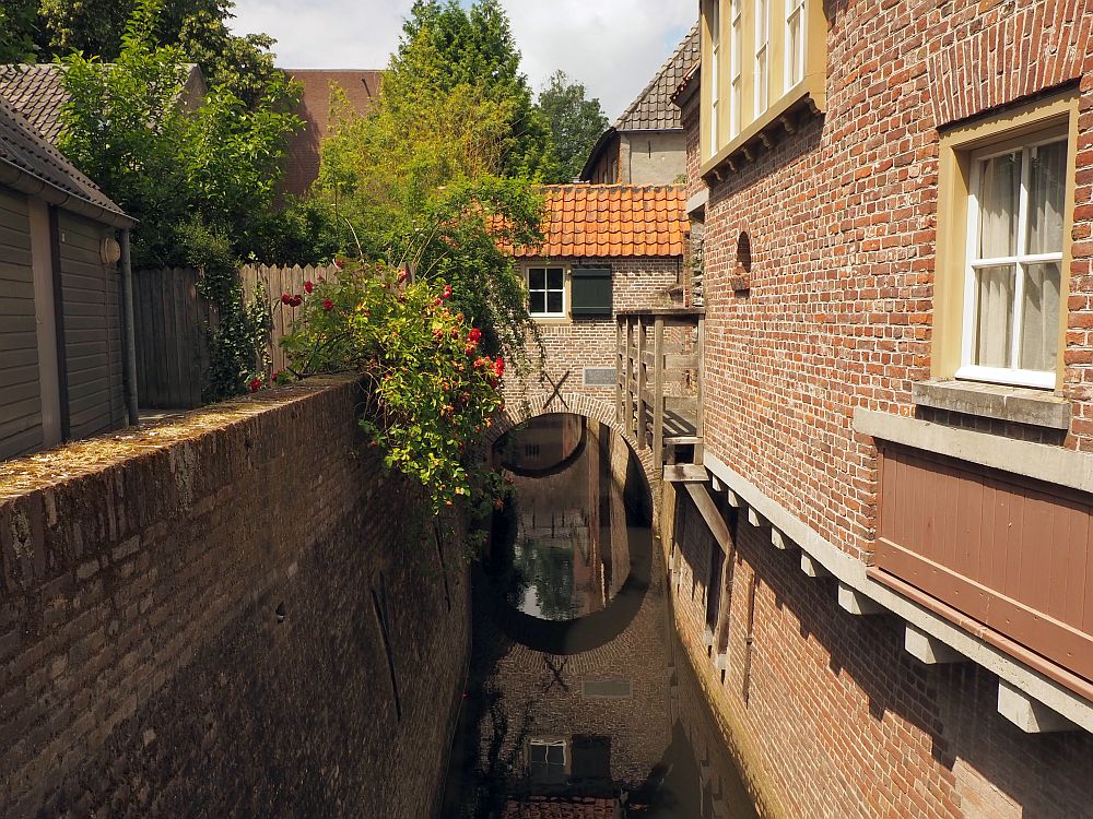 A canal-like stream down the middle going under an archway at the back of the view. Brick walls on both sides. The right side has a house above the wall, with windows on the wall. The left side has a walkway behind the wall. The archway at the back has a house on it, but only quite small from the looks of it: just one window above the stream.