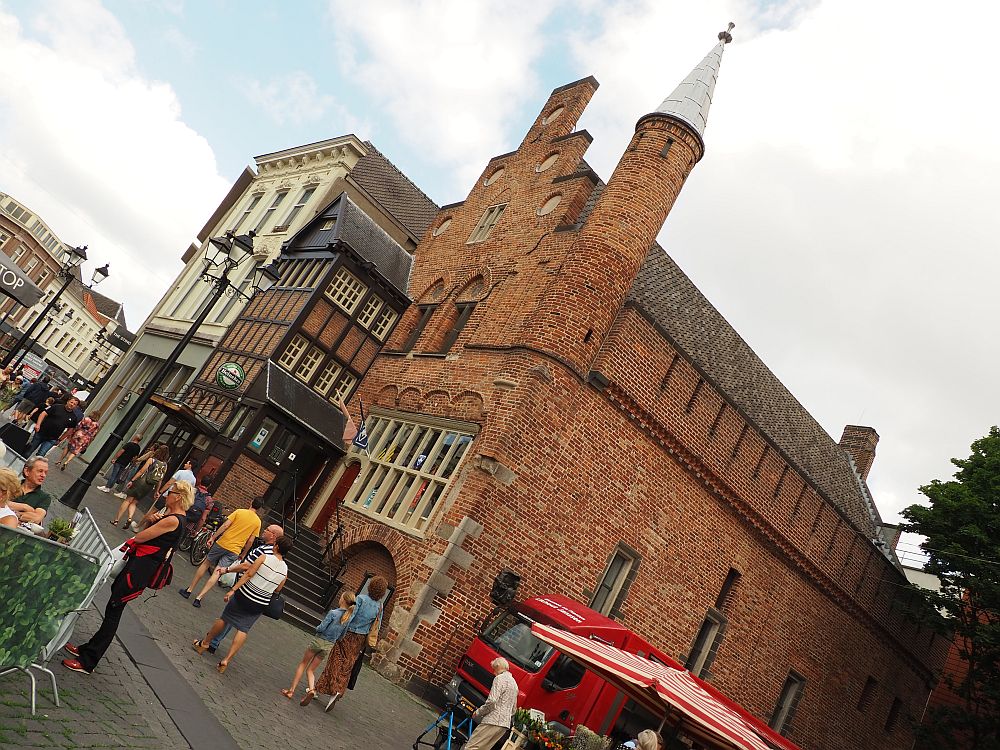 The building is red-brick with a stepped gable and otherwise a very simple front. On its corner is a round turret with a pointed roof. The photo was taken at an angle so the turret is pointing to the right top corner of the photo. 