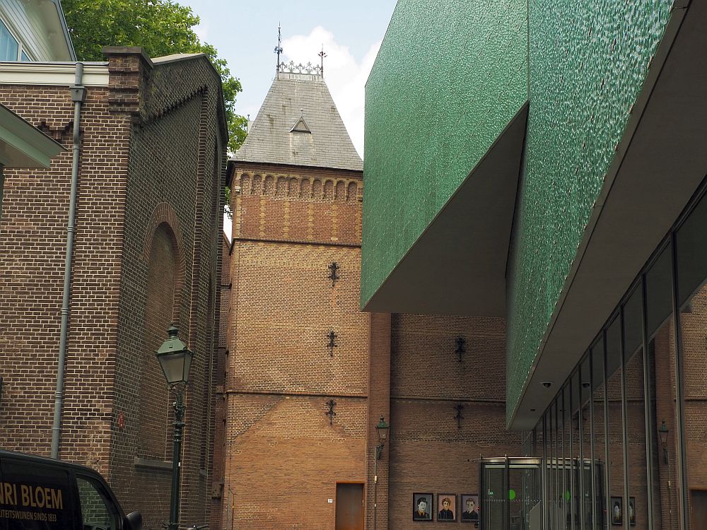 Looking down a narrow road. On the right is the museum: a green, windowless, massive wall above the ground floor, which is glass-sided. On the right, a brick building (the back of the synagogue). Ahead, a brick tower.