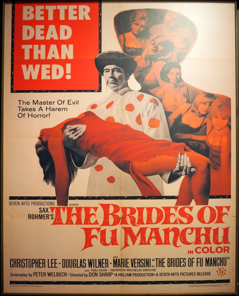 In the center is an image of the Fu Manchu character, looking at the camera, wearing a white robe with red polka dots. He has a mustache that hangs long and thin on either side of his mouth. He wears a wide-brimmed hat. He carries a woman under her back with one hand and under her knees with the other. She is dressed all in red, including red tights, and appears to be unconscious, with her ams and head dangling. Behind is an image of a group of women, all white, mostly dressed in bras. They look at the camera but do not appear to be afraid, but rather alluring. The text at the top says: Better dead than wed! The Master of Evil Takes A Harem of Horror! Below the image it says: Sax Rohmer's The Brides of Fu Manchu in Color, followed by a list of the main actors: Christopher Lee, Douglas Wilner, Marie Versini.