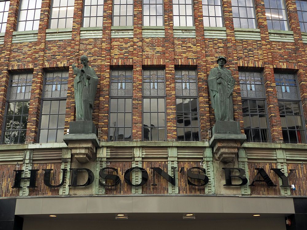 Along the bottom of the picture is the name of the store: Hudson's Bay. Above the lettering are two robed human figures (saints?). Three vertical windows between them and two on each side each have stained glass in their upper panes.