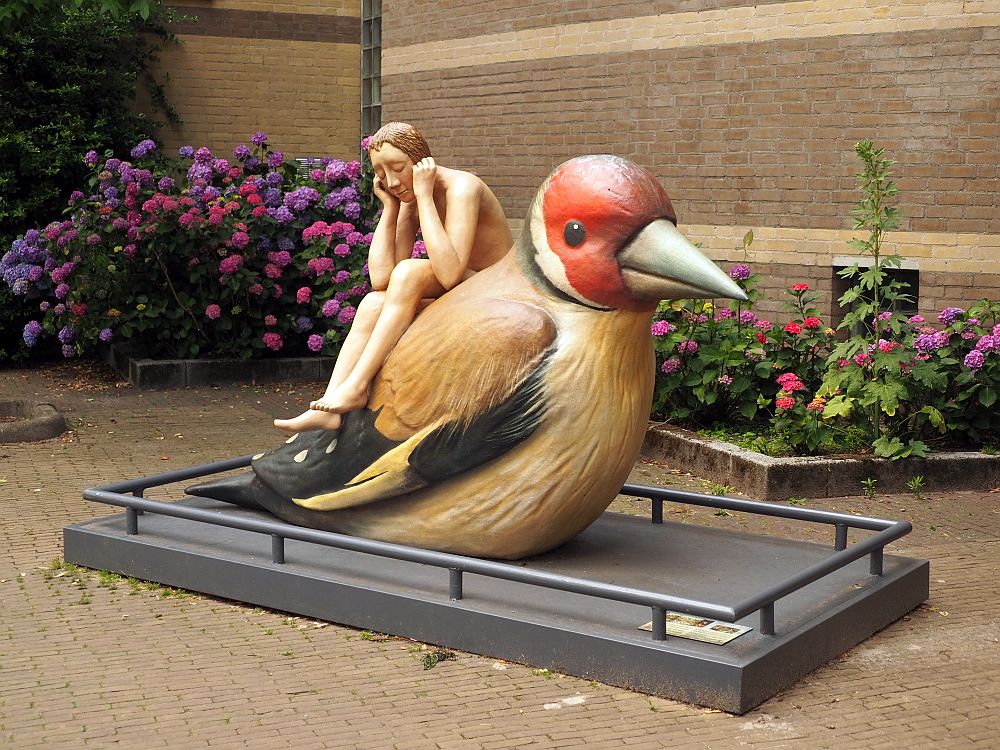 The person in the sculpture is a naked young man sitting on top of a bird's back. The man sits with his feet dangling; he leans forward so his elbows are on his knees and his hands are by his ears. The bird is brown with a red head, a black tail with white polka dots and a a brown beak.