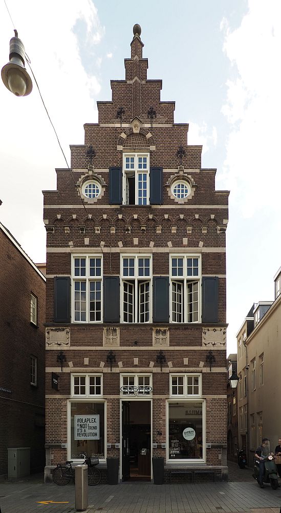 A tall, narrow red-brick building with a step gable. It is three stories tall. On the ground floor, a doorway in the middle and a window on either side of it. Three windows on the story above that. On the top story is one central rectangular window (like the others but smaller) flanked by two round windows. 