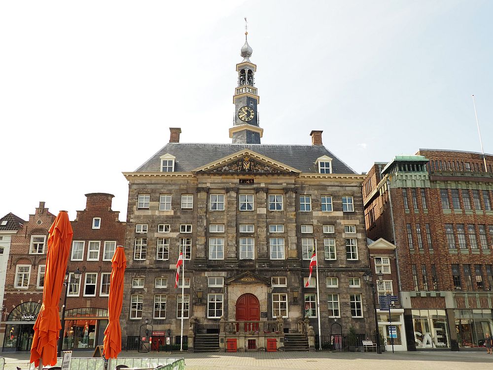 The building is quite large: it looks like it is 4 stories tall. The entry is exactly in the middle and has a red door up a flight of stairs on left and right. Four windows on either side of the entry. The story above that has 9 windoes across, so does the story above that, only they are smaller. The roof is above that and shows two dormer windows. The facade looks like it is some sort of stone and has flat pillar-like verticals around the central 6 windows and the door below. On top of the building is a tall narrow tower with a clock and, above that, a bell tower.