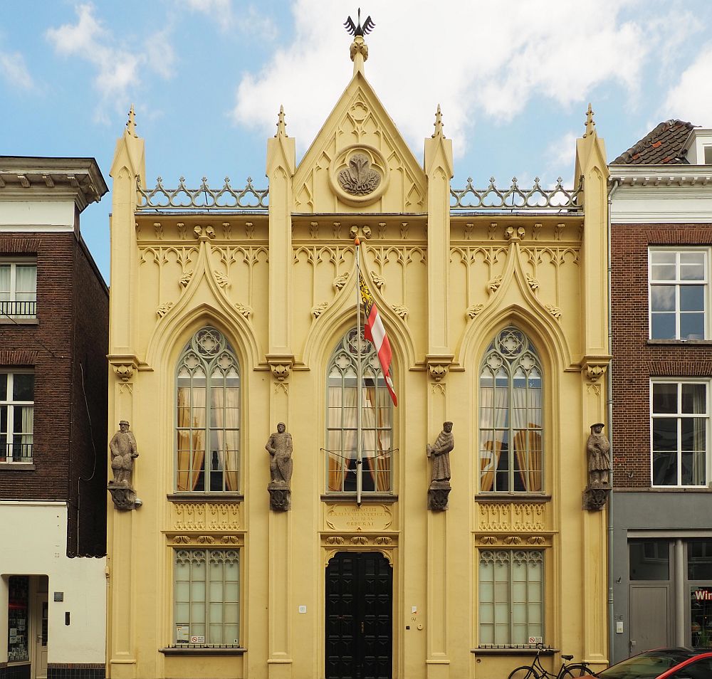 The building has a yellowish plaster with lots of decorative detail. It is two stories tall, with a door in the middle on the ground floor, flanked by one window on either side. On the upper story are three windows in gothic arch shape. The plaster work continues the gothic arch patterning above the windows. Four human figures (saints, maybe?) flank the three windows on the upper story. The roof has a small triangular point above the center window. A decorative medalion is in the center of the triangle and on its top point is a sculpture of a swan.