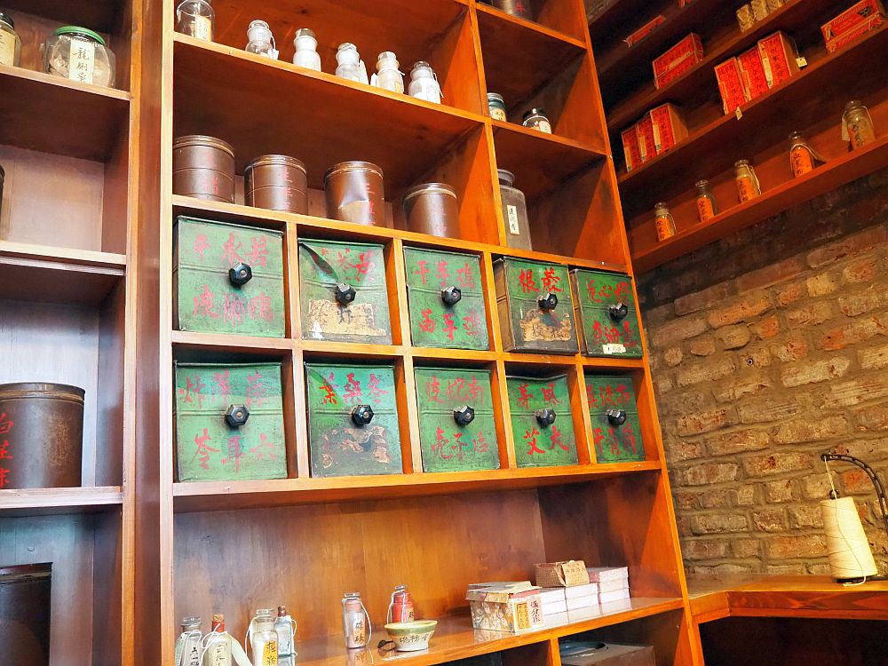 Neat brown wooden shelving, divided in squares and rectangular shelves. The middle ones are 5 squares across, each with a small bin inside it. The bins all have a round know in the center and are painted green with red Chinese lettering. Above and below, various other container: jars, pots and boxes, neatly spaced on the shelves.